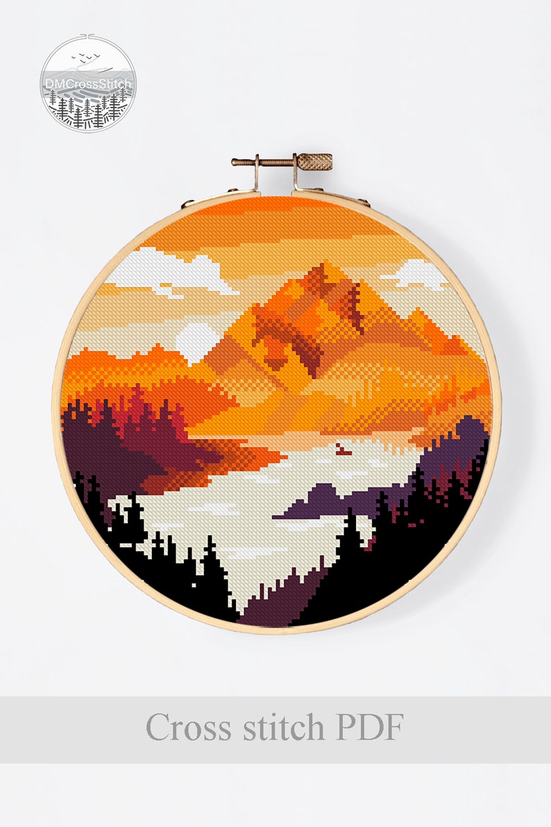 Op de afbeelding: Kruissteekpatroon van een berglandschap met een zonsondergangshemel. De bergen zijn oranje en bruin, met een meer op de voorgrond. De lucht is oranje en geel, met witte wolken. Het patroon is in een ronde borduurring.
