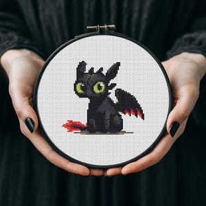 May include: A black cross-stitch of a dragon with green eyes and red wing accents, set in a round embroidery hoop. The dragon is sitting on a white background. The hoop is held by a person.