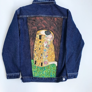 May include: A blue denim jacket with a painting of Gustav Klimt's "The Kiss" on the back. The painting features two figures in a golden embrace against a brown background.