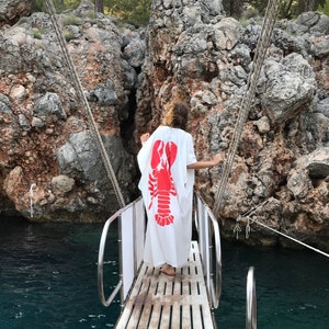 May include: A white caftan with a large red lobster design is worn by a person standing on a wooden dock. The dock leads to a boat, with a backdrop of rocky cliffs and water. The scene is outdoors, with trees visible.