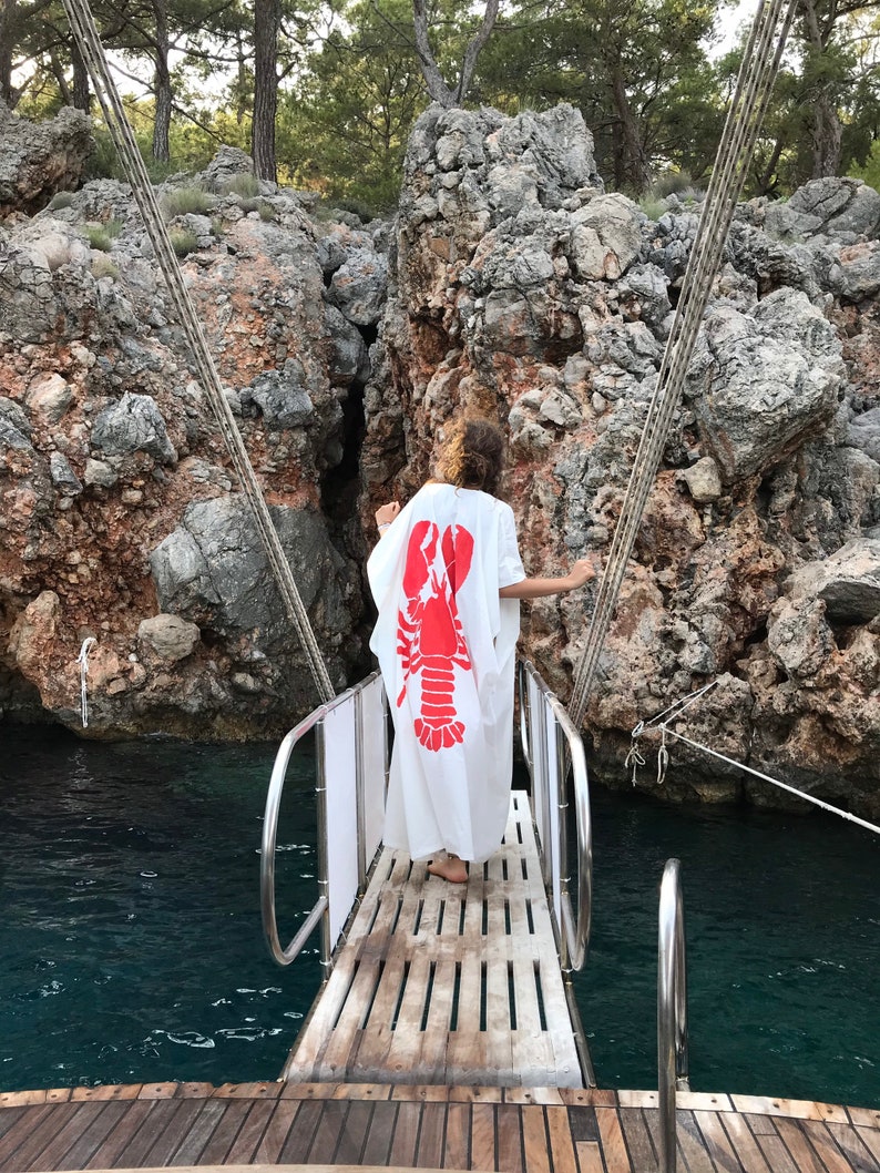 May include: A white caftan with a large red lobster design is worn by a person standing on a wooden dock. The dock leads to a boat, with a backdrop of rocky cliffs and water. The scene is outdoors, with trees visible.