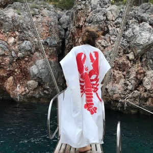 May include: A white, flowing garment with a large red lobster design on the back. The person is standing on a wooden boat deck, with a rocky cliff and water in the background. The garment appears to be a beach cover-up or robe.