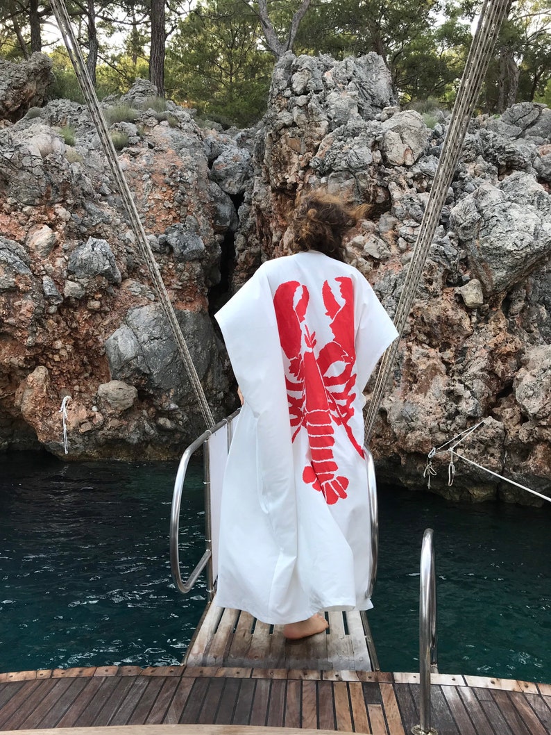May include: A white, flowing garment with a large red lobster design on the back. The person is standing on a wooden boat deck, with a rocky cliff and water in the background. The garment appears to be a beach cover-up or robe.