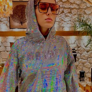 K&ouml;nnte beinhalten: Ein silberner, schimmernder Hoodie mit geometrischem Muster. Der Hoodie hat eine Kapuze mit Kordelzug und lange &Auml;rmel. Die Person, die den Hoodie tr&auml;gt, tr&auml;gt braune, quadratische Sonnenbrillen.