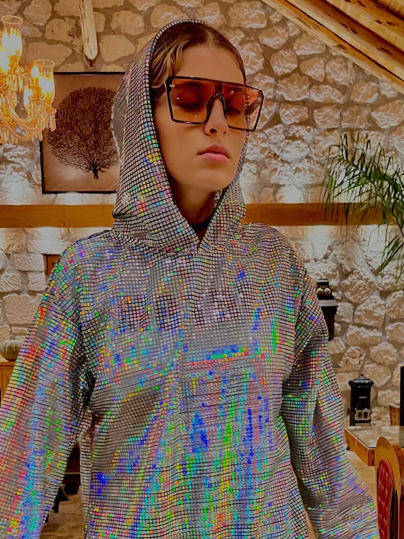 K&ouml;nnte beinhalten: Ein silberner, schimmernder Hoodie mit geometrischem Muster. Der Hoodie hat eine Kapuze mit Kordelzug und lange &Auml;rmel. Die Person, die den Hoodie tr&auml;gt, tr&auml;gt braune, quadratische Sonnenbrillen.