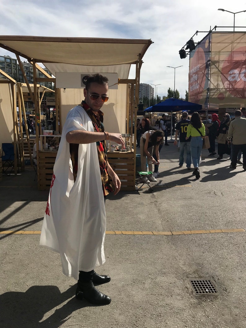 May include: A person wearing a white robe with red text, a patterned shirt, sunglasses, and black boots stands in an outdoor market setting. The background includes market stalls and other people.