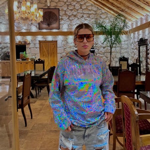 K&ouml;nnte beinhalten: Eine Person tr&auml;gt ein silbernes, schimmerndes Hoodie mit glitzernder Textur und zerrissene Jeans-Shorts. Sie tr&auml;gt eine gro&szlig;e, schwarze, quadratische Sonnenbrille und eine silberne Kettenkette.