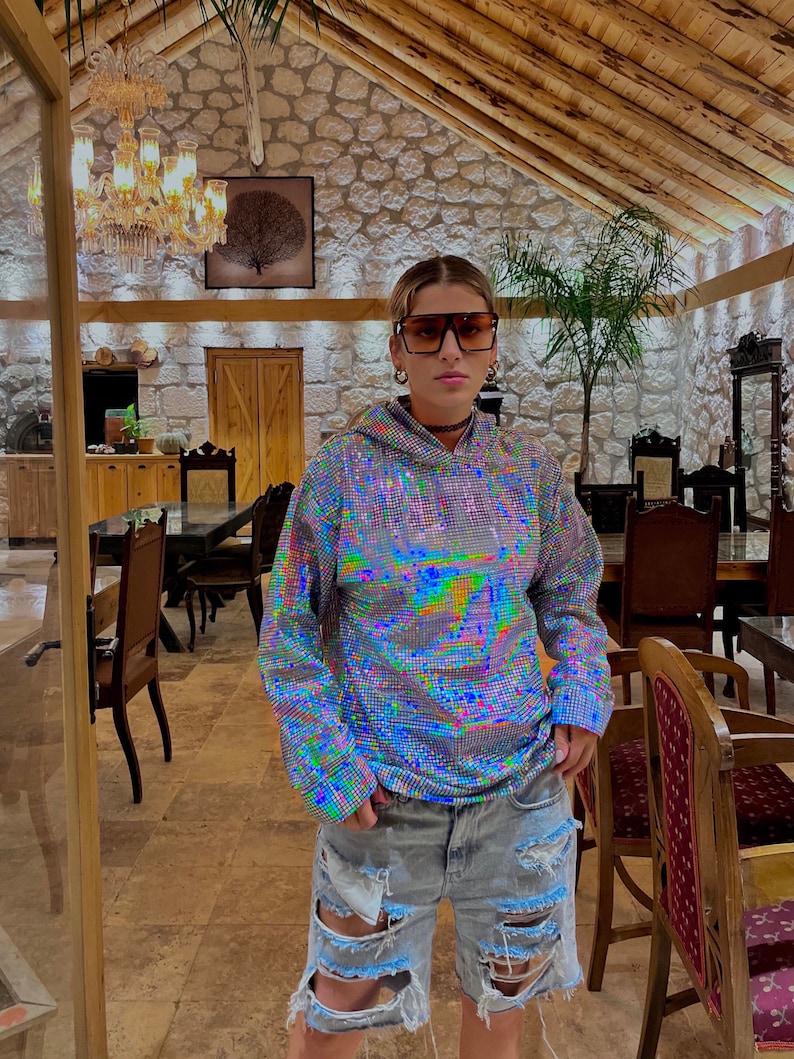 K&ouml;nnte beinhalten: Eine Person tr&auml;gt ein silbernes, schimmerndes Hoodie mit glitzernder Textur und zerrissene Jeans-Shorts. Sie tr&auml;gt eine gro&szlig;e, schwarze, quadratische Sonnenbrille und eine silberne Kettenkette.