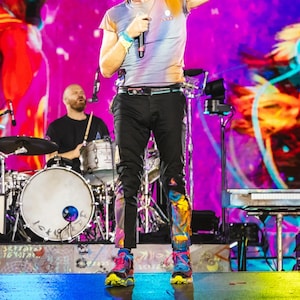 May include: A short-sleeved crew neck t-shirt with a blue and orange gradient pattern, paired with black trousers featuring colorful paint-splatter designs on the lower legs and bright blue and pink athletic sneakers with yellow soles.