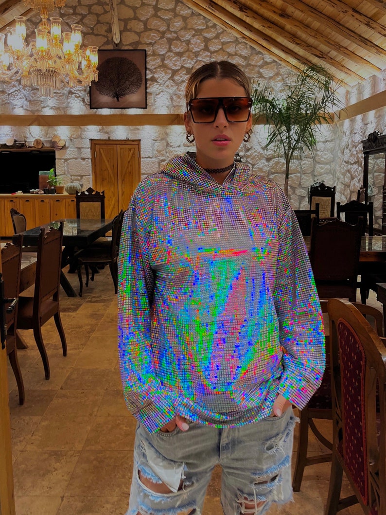 K&ouml;nnte beinhalten: Eine Person tr&auml;gt einen silbernen, schimmernden Hoodie mit einer schwarzen Halskette und eine blaue Jeans mit Rissen. Die Person tr&auml;gt eine Sonnenbrille und steht in einem Raum mit Holzbalken und Steinw&auml;nden.