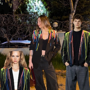 Puede incluir: Un blazer negro con patrones de pintura de goteo en colores neón vibrantes como rosa, amarillo, azul y naranja en los hombros y solapas. La chaqueta tiene mangas largas, cuello de solapa y un corte estructurado con un diseño de salpicaduras.