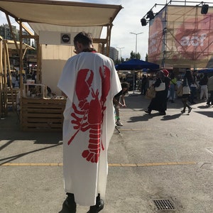 May include: A white robe with a large red lobster graphic on the back. The robe is worn by a person standing in an outdoor market setting. The image is taken from behind, showing the robe's design and the surrounding environment.