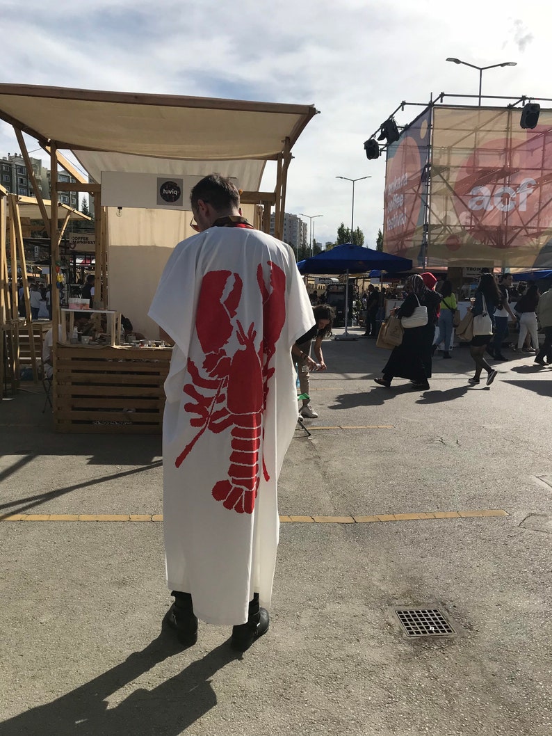 May include: A white robe with a large red lobster graphic on the back. The robe is worn by a person standing in an outdoor market setting. The image is taken from behind, showing the robe's design and the surrounding environment.