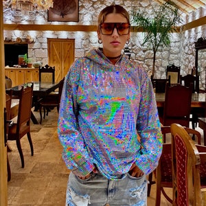 K&ouml;nnte beinhalten: Eine Person tr&auml;gt ein silbernes Pailletten-Hoodie mit einem regenbogenfarbenen, schillernden Muster und zerrissene Jeans-Shorts. Die Person steht in einem Raum mit einer Holzdecke und Steinw&auml;nden.