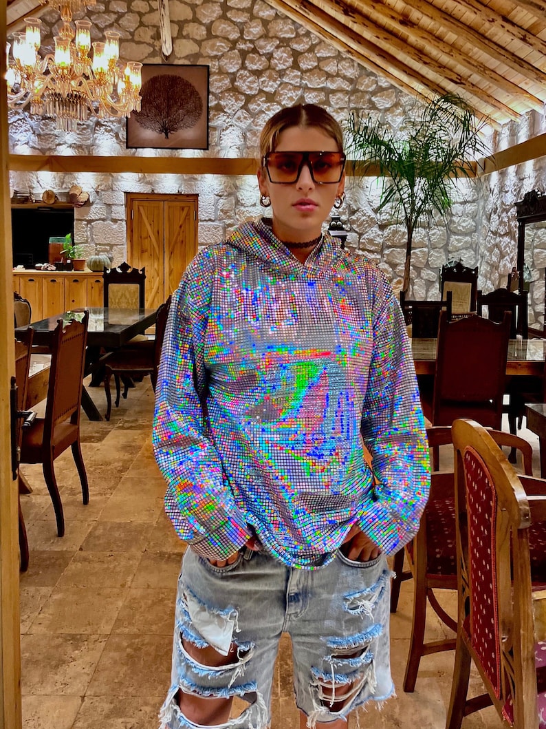K&ouml;nnte beinhalten: Eine Person tr&auml;gt ein silbernes Pailletten-Hoodie mit einem regenbogenfarbenen, schillernden Muster und zerrissene Jeans-Shorts. Die Person steht in einem Raum mit einer Holzdecke und Steinw&auml;nden.