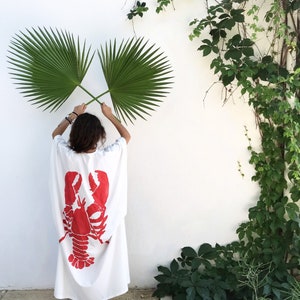 May include: A white caftan dress with a large red lobster graphic on the back. The person is holding two green palm leaves in front of a white wall with green vines.