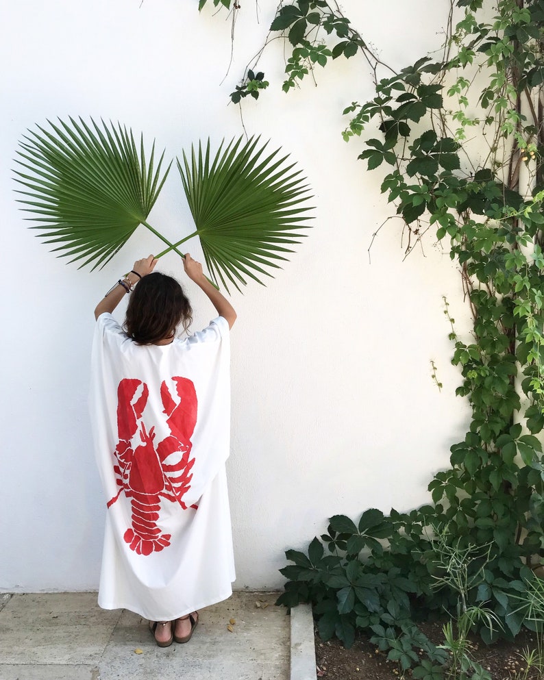 May include: A white caftan dress with a large red lobster graphic on the back. The person is holding two green palm leaves in front of a white wall with green vines.