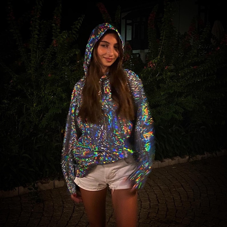 K&ouml;nnte beinhalten: Eine Frau tr&auml;gt einen silbernen, regenbogenfarbenen Hoodie mit Kordelzug. Der Hoodie hat eine reflektierende, holografische Oberfl&auml;che.