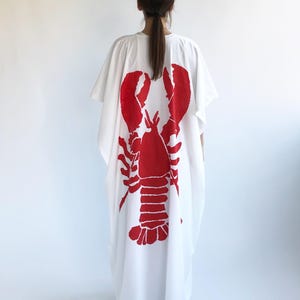 May include: White caftan dress featuring a large red lobster graphic on the back. The loose-fitting garment has short sleeves and a relaxed silhouette. The lobster design is a bold, eye-catching print.