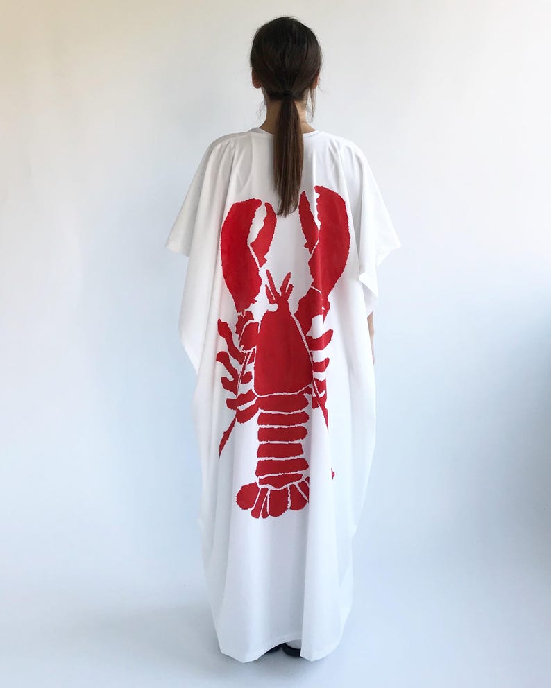 May include: White caftan dress featuring a large red lobster graphic on the back. The loose-fitting garment has short sleeves and a relaxed silhouette. The lobster design is a bold, eye-catching print.