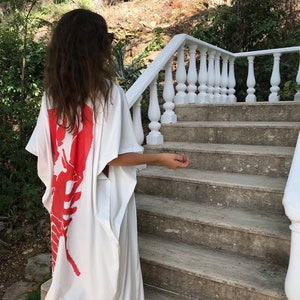 May include: A white caftan dress featuring a large red lobster design on the back. The dress has a loose, flowing silhouette. The person is standing on a stone staircase with a white railing, with greenery in the background.