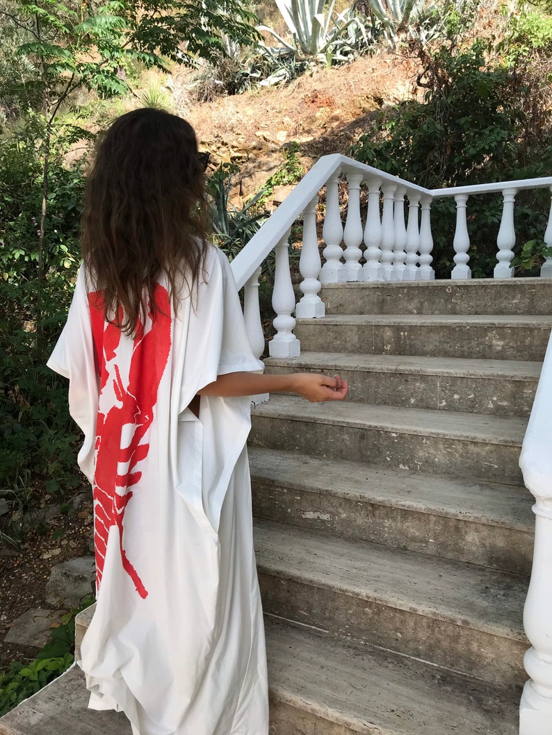 May include: A white caftan dress featuring a large red lobster design on the back. The dress has a loose, flowing silhouette. The person is standing on a stone staircase with a white railing, with greenery in the background.