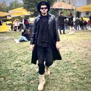 Puede incluir: Una persona que lleva un abrigo negro de cuero, pantalones negros y botas de color canela. Lleva una camisa negra y un sombrero negro con un collar de cadena dorada. La persona está de pie en un área cubierta de hierba con otras personas en el fondo.