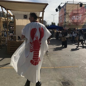 May include: A white kimono-style garment with a large red lobster graphic on the back. The garment is worn outdoors, with a person wearing it. The background includes market stalls and people.