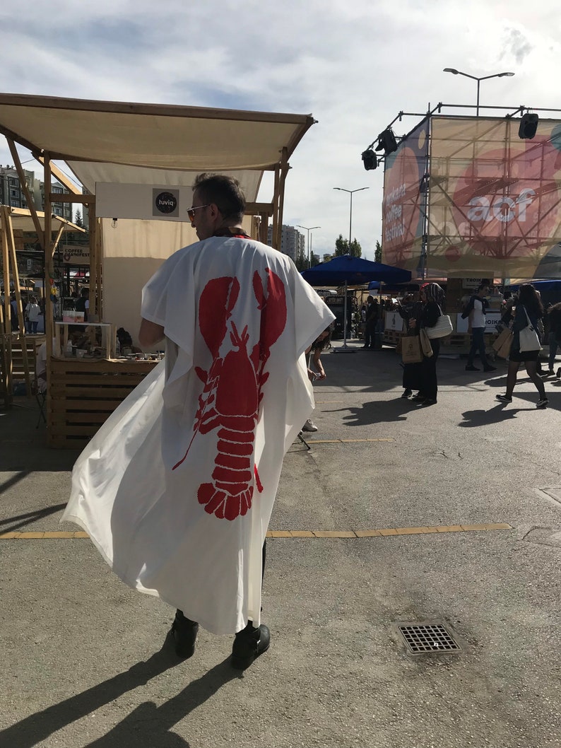 May include: A white kimono-style garment with a large red lobster graphic on the back. The garment is worn outdoors, with a person wearing it. The background includes market stalls and people.