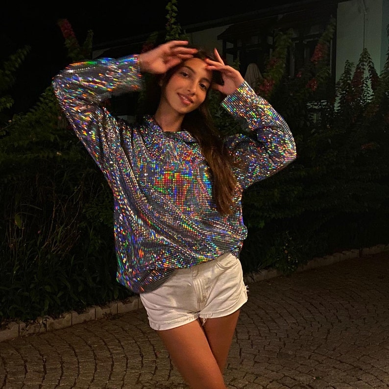 K&ouml;nnte beinhalten: Eine Frau tr&auml;gt ein silbernes, irisierendes, sequinenbesetztes Langarmhemd mit einem wei&szlig;en Hoodie und einer wei&szlig;en Jeans-Shorts.