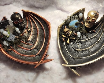May include: Two decorative Halloween-themed trays. One is black and copper, the other black and gold. Each features a skull, crystals, moss, and a bat wing design. One has a tombstone that says RIP.