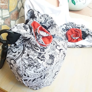 Könnte beinhalten: Eine Tasche aus schwarz-weißem Paisley-Stoff mit einem schwarzen runden Griff und einer auffälligen Stoffblüte in Rostorange. Daneben liegt ein passendes Stoffaccessoire mit einer kleineren rostorangen Stoffblüte und schwarzem Rand.