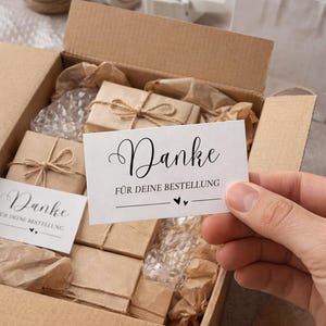 Könnte beinhalten: Ein Karton gefüllt mit verpackten Geschenken, Luftpolsterfolie und Packpapier. Eine Hand hält eine weiße Karte mit dem Wort "Danke" und "Für deine Bestellung" darauf. Garn, Klebeband und eine weiße Einkaufstasche sind ebenfalls zu sehen.