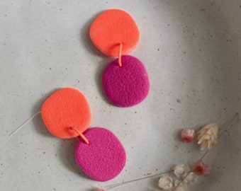 May include: Two pairs of polymer clay earrings. One pair is pink and orange, the other is orange and pink. The earrings are displayed on a white ceramic bowl.