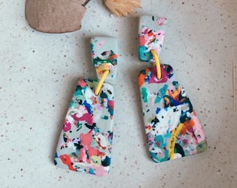 Puede incluir: Un par de pendientes colgantes abstractos y coloridos con una base gris y detalles amarillos. Los pendientes están hechos de arcilla polimérica y tienen un diseño de pintura salpicada.