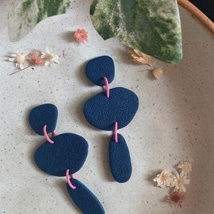 Puede incluir: Un par de pendientes de declaración azules y rosas con un diseño geométrico. Los pendientes están hechos de arcilla polimérica y tienen un acabado mate.