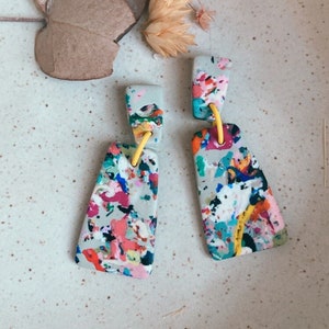 Puede incluir: Un par de pendientes colgantes abstractos y coloridos con una base gris y detalles amarillos. Los pendientes están hechos de arcilla polimérica y tienen un diseño de pintura salpicada.