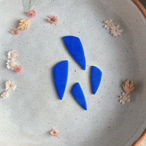 以下が含まれることがあります： 白い陶器の皿の上に、青い陶器の三角形が4つ置かれ、その周りにドライフラワーが散らばっている。