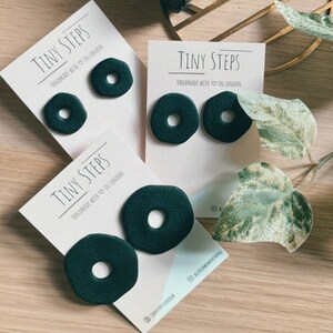 May include: Dark green donut-shaped earrings on display cards with the text "TINY STEPS HANDMADE WITH IN LONDON". A wrapped gift box tied with twine sits above the earrings. The earrings are approximately 2.5 cm in diameter.