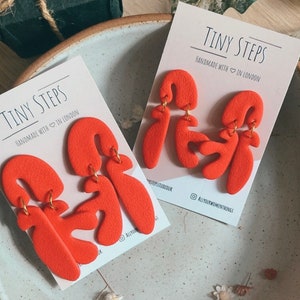 May include: A pair of orange abstract shaped earrings with gold hoops. The earrings are displayed on a white card with the text "Tiny Steps Handmade with in London".