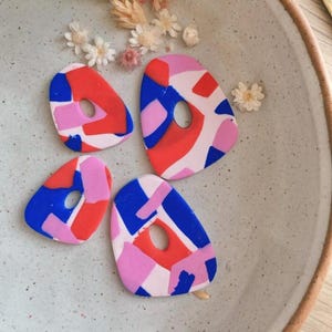 May include: Three colourful abstract geometric shapes made of polymer clay. The shapes are pink, blue and red with white accents. The shapes are arranged on a white ceramic plate.