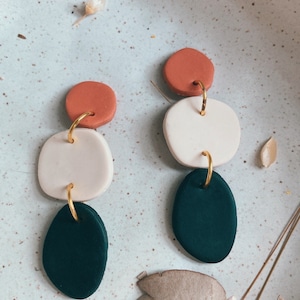 Puede incluir: Un par de pendientes de arcilla polimérica hechos a mano. Cada pendiente presenta tres formas geométricas: un círculo color óxido, un óvalo blanco y una lágrima verde oscuro, conectados por aros dorados. Los pendientes se exhiben sobre una superficie azul claro moteada.
