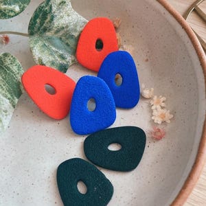 Puede incluir: Cuatro pendientes geométricos coloridos con un agujero en el centro. Los pendientes son en tonos de rojo, azul y verde.