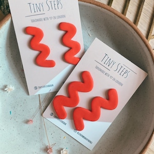 May include: Two pairs of red polymer clay earrings shaped like a zig-zag pattern. The earrings are displayed on a white card with the text "Tiny Steps Handmade with  in London" and the social media handle "@tinystepsstudio.uk".