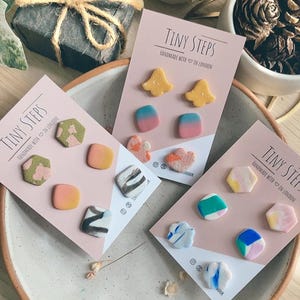 May include: Three sets of handmade polymer clay earrings displayed on cards. The first set features a yellow bell shape, the second set features a black and white abstract design, and the third set features a blue and green abstract design. Each card has the text "Tiny Steps" and "Handmade with in London".