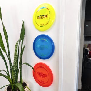 May include: Three colorful disc golf discs are displayed on a white wall. The top disc is yellow with the word "NUKE" in black. The middle disc is blue with "BUZZZ SS" in blue. The bottom disc is red with "ZONE OS" in red.