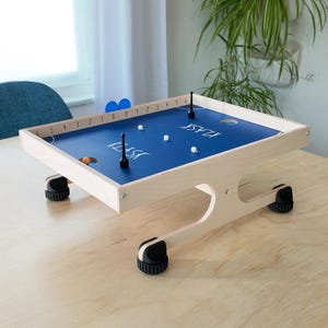 May include: A wooden Klask game with a blue playing surface. The game features a white wooden frame, black and white game pieces, and the word "KLASK" printed on the surface. The game is set on a light wood table.
