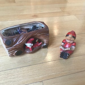 May include: A ceramic planter shaped like a tree trunk with a small elf figurine inside. A small ceramic figurine of a girl in a red dress is sitting on the floor, reading a book.