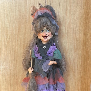 May include: A handmade witch puppet with long gray hair, blue eyes, and a crooked smile. The witch wears a purple and green dress with a black shawl and a matching hat. She holds a broom, perfect for Halloween.