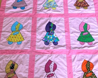 Puede incluir: Una colcha rosa con nueve cuadrados, cada uno con una muñeca aplicada diferente. Las muñecas llevan todas vestidos con diferentes patrones y colores, incluyendo amarillo, azul, verde, rosa y rojo. Las muñecas tienen ojos negros y cabello negro.